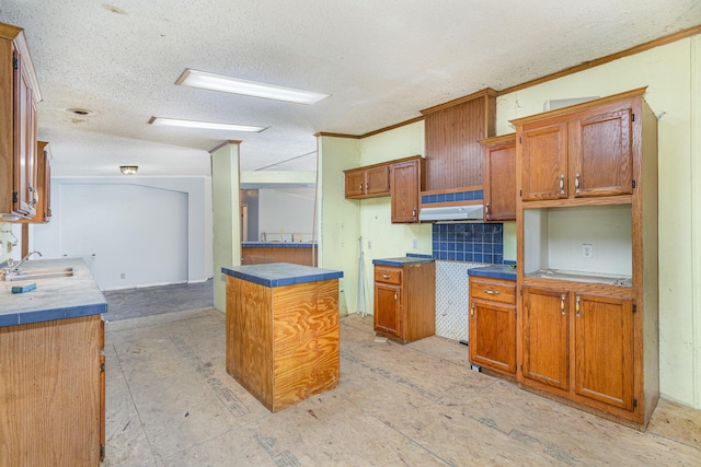 kitchen featuring brown cabinetry, a sink, under cabinet range hood, a textured ceiling, and crown molding