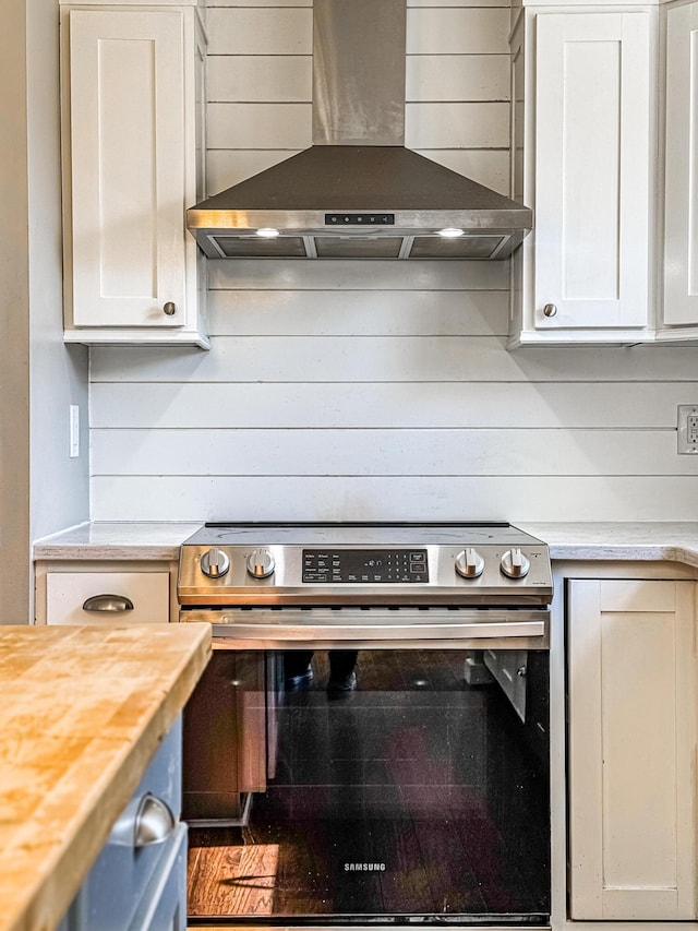 kitchen featuring wooden walls, butcher block counters, stainless steel electric range, white cabinets, and wall chimney exhaust hood