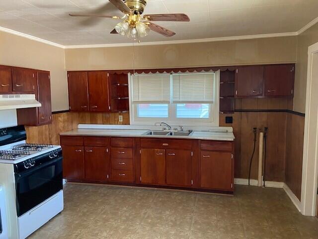 kitchen with wooden walls, ceiling fan, ornamental molding, range with gas stovetop, and sink