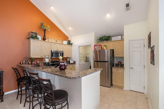 kitchen featuring appliances with stainless steel finishes, washing machine and clothes dryer, light brown cabinetry, kitchen peninsula, and vaulted ceiling