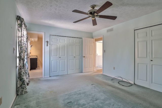 unfurnished bedroom featuring ensuite bath, light colored carpet, a textured ceiling, two closets, and ceiling fan