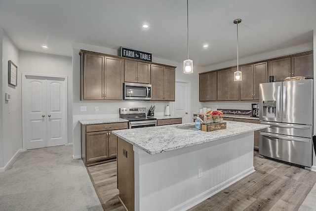 kitchen with light stone counters, hanging light fixtures, light hardwood / wood-style flooring, stainless steel appliances, and a kitchen island with sink