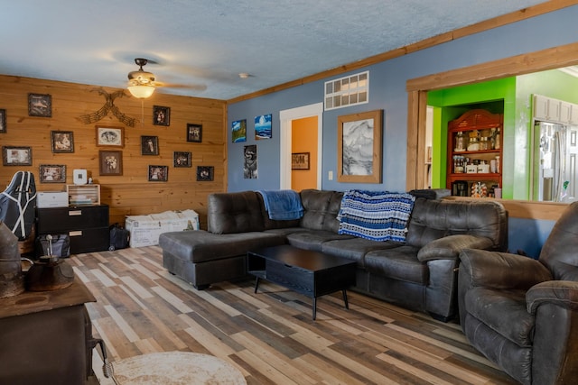 living room with ceiling fan, wood-type flooring, ornamental molding, a textured ceiling, and wood walls