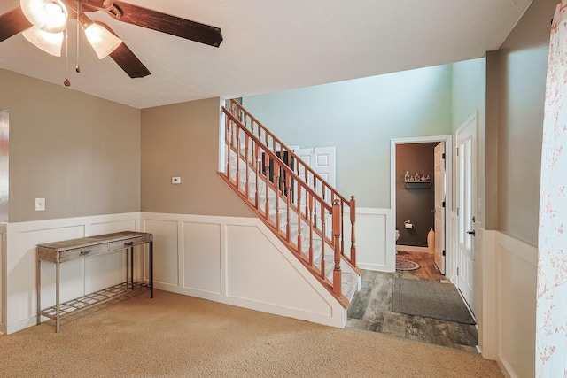 stairway featuring carpet, wainscoting, a decorative wall, and ceiling fan