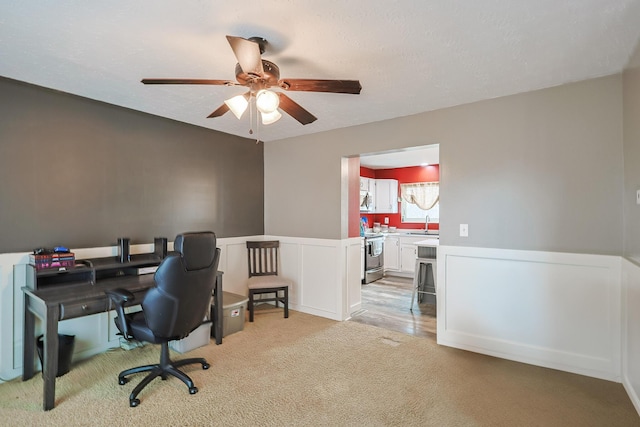 home office featuring ceiling fan, a textured ceiling, a decorative wall, light colored carpet, and wainscoting