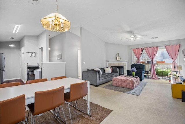 carpeted dining space with lofted ceiling, visible vents, a tiled fireplace, and ceiling fan with notable chandelier