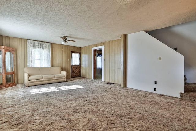 unfurnished living room featuring stairway, ceiling fan, wood walls, a textured ceiling, and carpet flooring