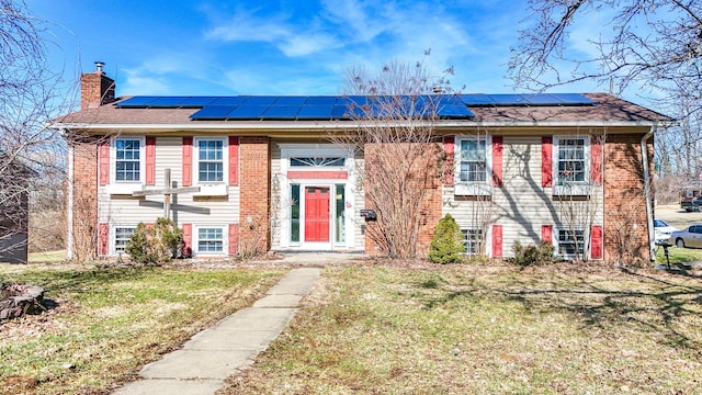 split foyer home featuring brick siding, a chimney, a front yard, and roof mounted solar panels