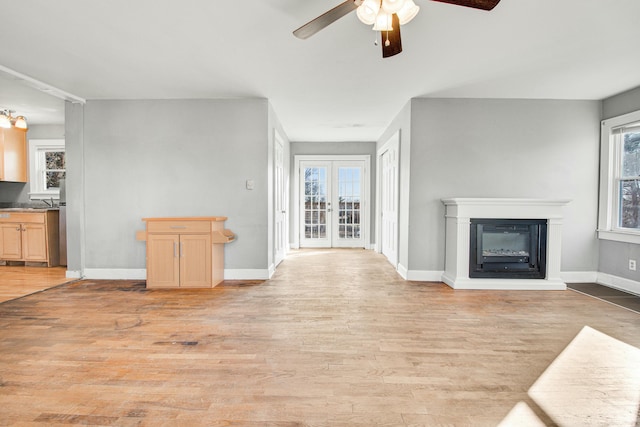 unfurnished living room featuring french doors, light wood finished floors, a ceiling fan, a glass covered fireplace, and baseboards