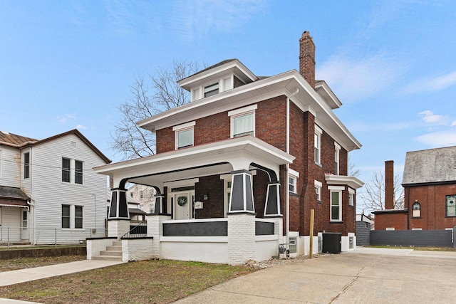 american foursquare style home with brick siding, a chimney, covered porch, central AC, and fence