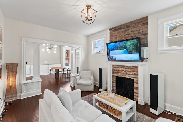 living area with a stone fireplace, a notable chandelier, dark wood-type flooring, baseboards, and ornate columns