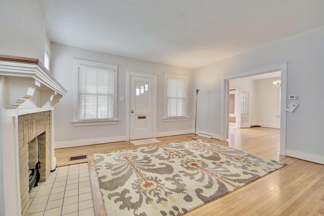 foyer entrance with baseboards, a brick fireplace, visible vents, and light wood-style floors