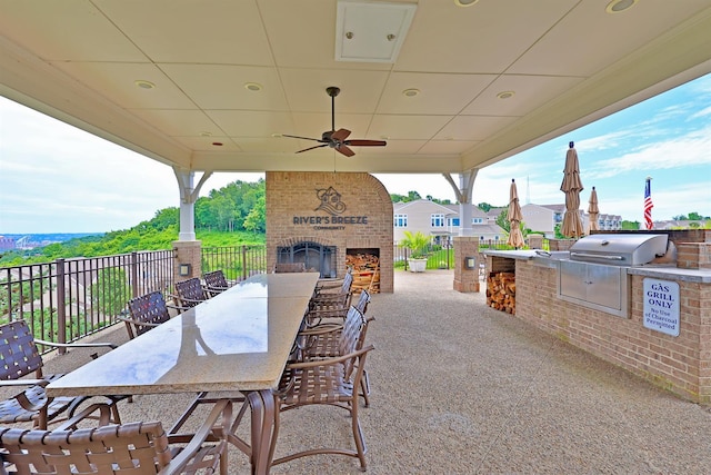 view of patio / terrace featuring fence, an outdoor kitchen, area for grilling, an outdoor brick fireplace, and ceiling fan