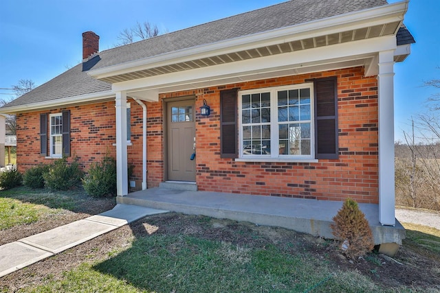 entrance to property featuring covered porch, brick siding, roof with shingles, and a chimney