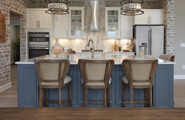 kitchen featuring a kitchen island with sink, light countertops, appliances with stainless steel finishes, wall chimney range hood, and a chandelier