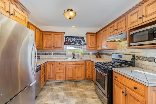 kitchen with tile countertops, brown cabinetry, a sink, under cabinet range hood, and appliances with stainless steel finishes