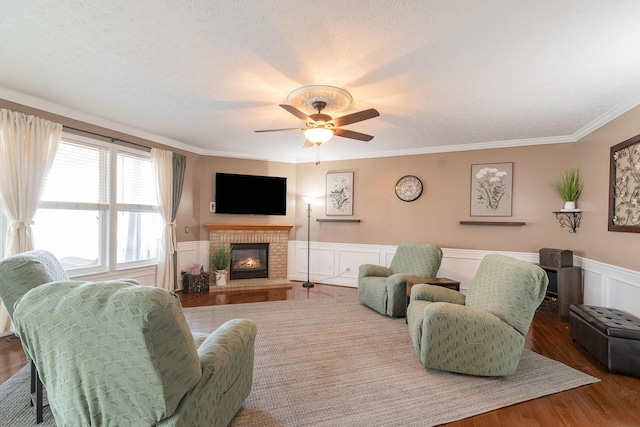 living room with wood finished floors, a wainscoted wall, ornamental molding, ceiling fan, and a brick fireplace