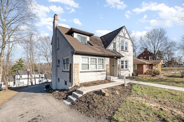 view of front facade with driveway, brick siding, roof with shingles, and a chimney