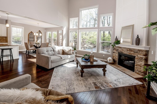 living room featuring an inviting chandelier, a towering ceiling, a stone fireplace, dark wood-style flooring, and baseboards