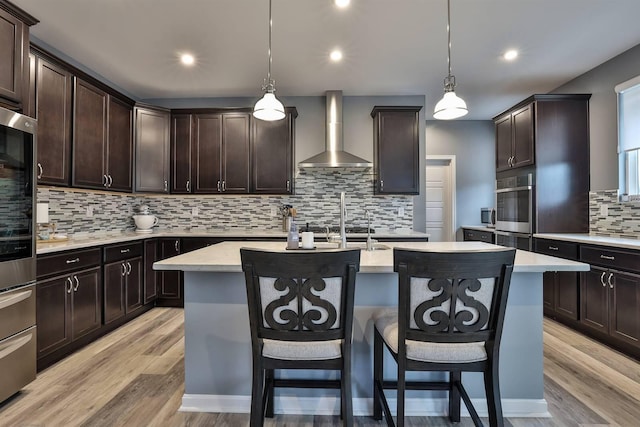 kitchen with wall chimney range hood, an island with sink, dark brown cabinetry, stainless steel appliances, and decorative backsplash