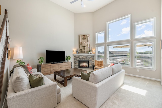 living area featuring baseboards, light carpet, a ceiling fan, a stone fireplace, and a high ceiling