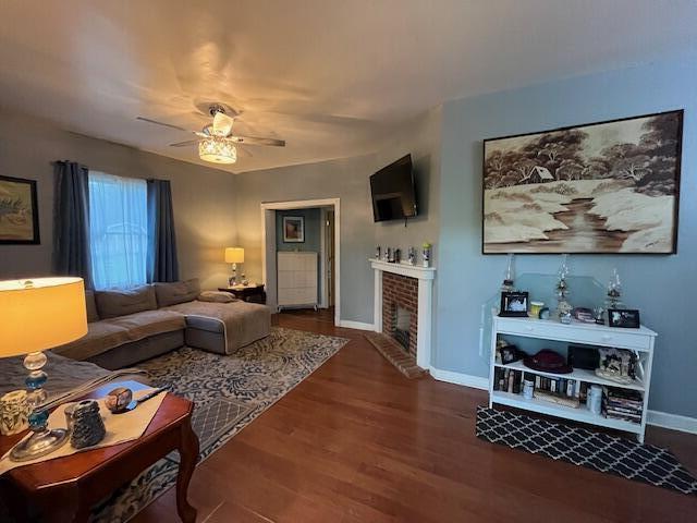 living area featuring baseboards, a ceiling fan, a brick fireplace, and wood finished floors