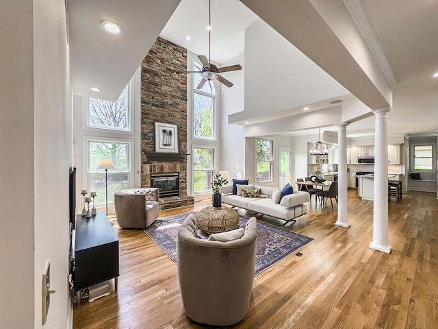 living room with a high ceiling, ceiling fan, a stone fireplace, light wood-style flooring, and ornate columns