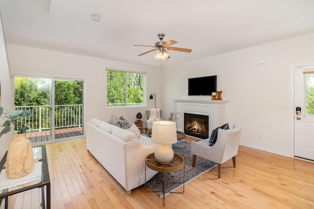 living room with light wood-type flooring, a ceiling fan, baseboards, and a glass covered fireplace