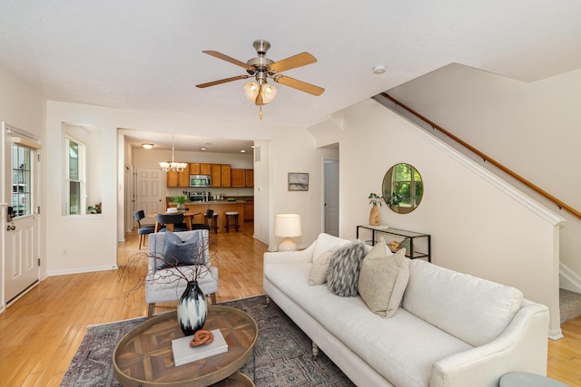 living area featuring baseboards, light wood finished floors, ceiling fan with notable chandelier, and stairs