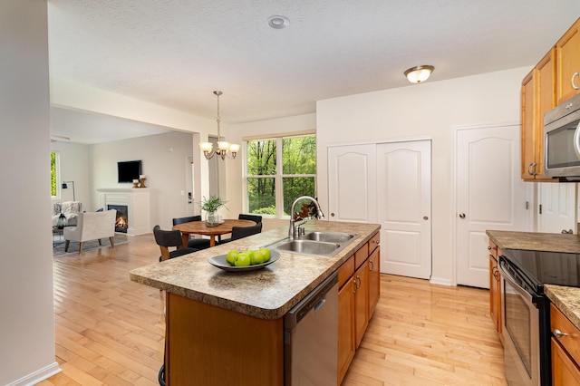 kitchen featuring a center island with sink, a warm lit fireplace, a sink, light wood-style flooring, and stainless steel appliances