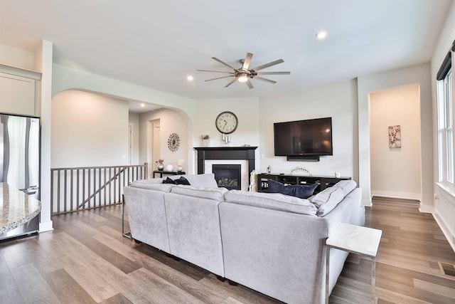 living area with arched walkways, a glass covered fireplace, a ceiling fan, baseboards, and wood finished floors