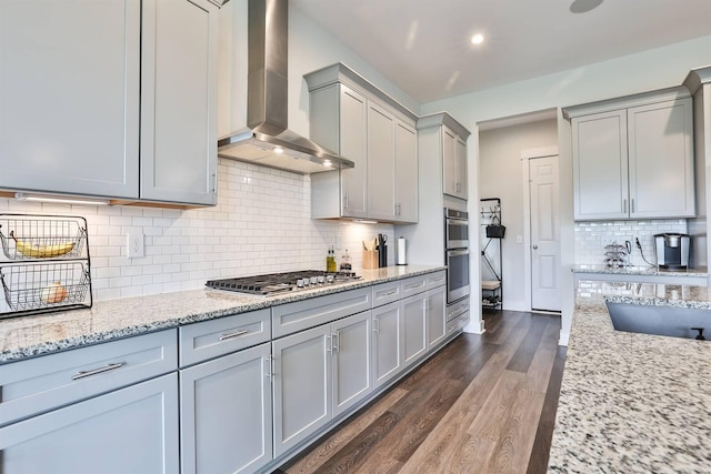 kitchen with decorative backsplash, gray cabinetry, dark wood-style floors, stainless steel appliances, and wall chimney exhaust hood