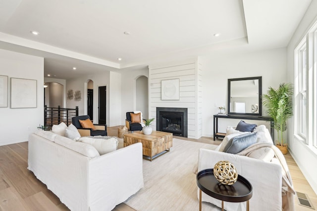 living room with a raised ceiling, arched walkways, light wood-style floors, recessed lighting, and a glass covered fireplace