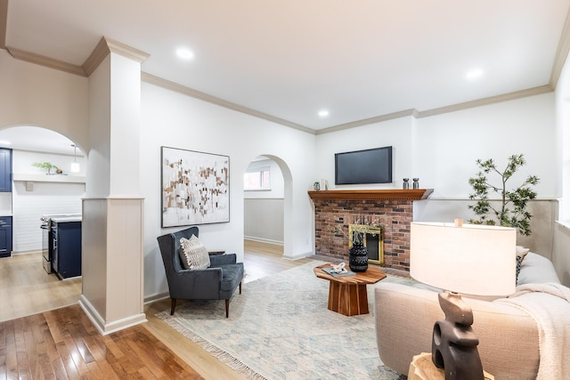 living area featuring light wood finished floors, a brick fireplace, crown molding, baseboards, and arched walkways