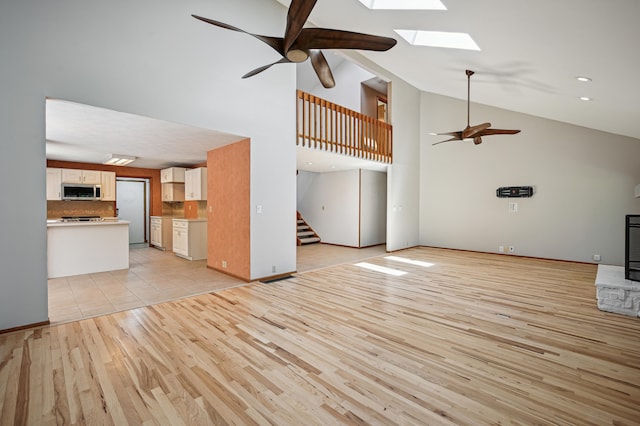unfurnished living room with a ceiling fan, a skylight, stairway, light wood finished floors, and high vaulted ceiling