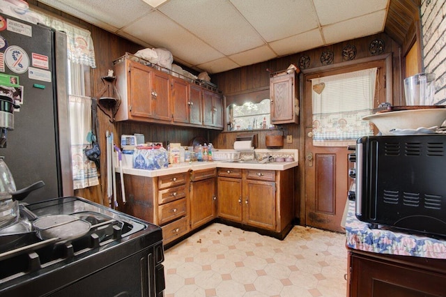 kitchen featuring light floors, wood walls, black range with gas stovetop, freestanding refrigerator, and brown cabinetry