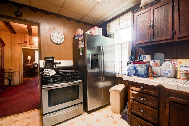 kitchen with appliances with stainless steel finishes, light floors, tile counters, a drop ceiling, and dark brown cabinets