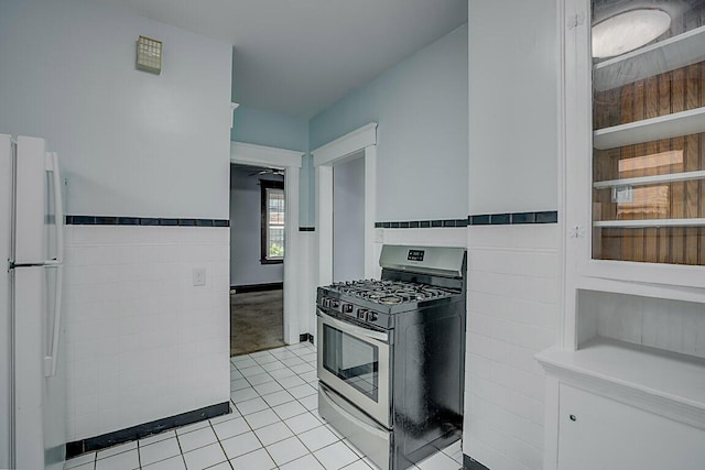 kitchen featuring stainless steel range with gas stovetop, light tile patterned floors, freestanding refrigerator, tile walls, and wainscoting