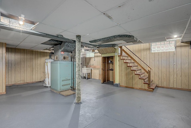 basement featuring a drop ceiling, water heater, wood walls, stairway, and a sink