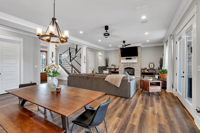 dining room with ceiling fan with notable chandelier, wood finished floors, stairway, crown molding, and a fireplace