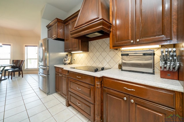 kitchen with premium range hood, light tile patterned flooring, stainless steel refrigerator, backsplash, and black electric cooktop