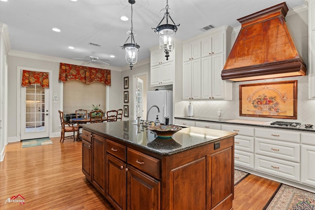 kitchen featuring white fridge, dark stone countertops, premium range hood, white cabinetry, and an island with sink