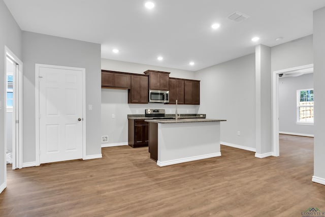 kitchen with light wood-type flooring, stainless steel appliances, a kitchen island with sink, and dark stone counters