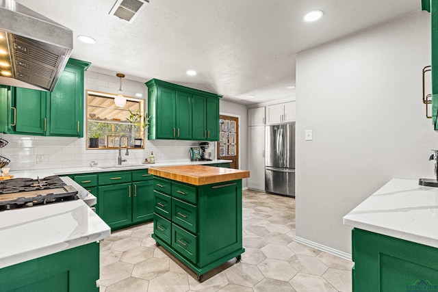 kitchen with backsplash, green cabinets, a sink, stainless steel fridge, and exhaust hood