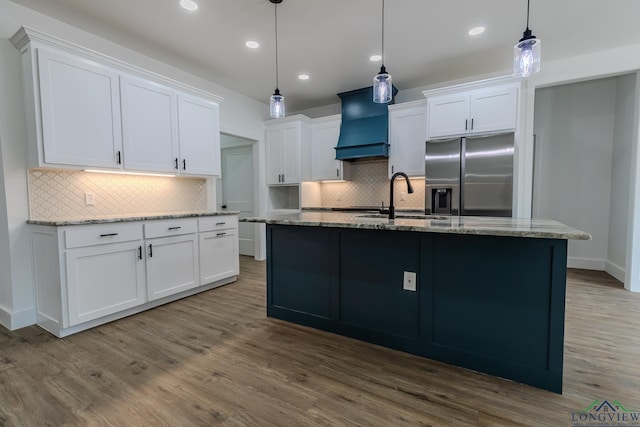 kitchen with white cabinets, hanging light fixtures, stainless steel fridge with ice dispenser, custom range hood, and a center island with sink