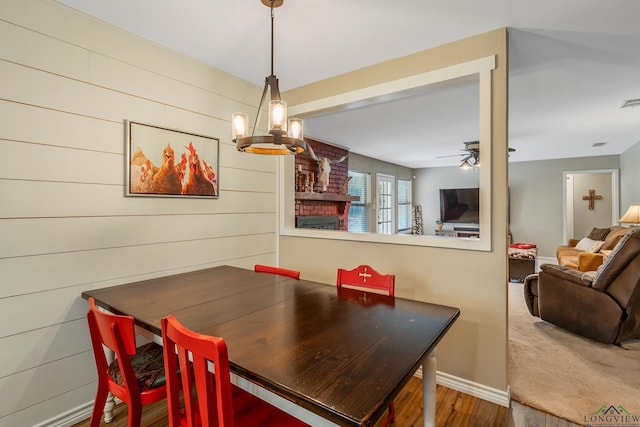 dining space featuring wood walls, a fireplace, ceiling fan with notable chandelier, and hardwood / wood-style flooring