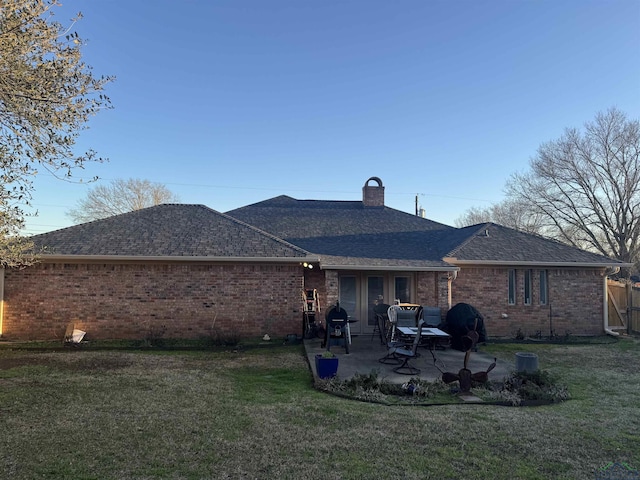 rear view of property with roof with shingles, brick siding, a patio, a chimney, and a lawn