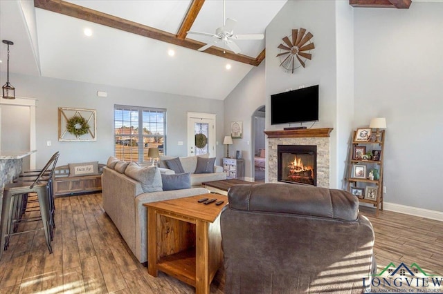 living room featuring ceiling fan, beam ceiling, hardwood / wood-style flooring, high vaulted ceiling, and a stone fireplace