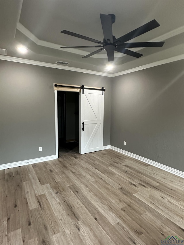 empty room featuring ceiling fan, a tray ceiling, light hardwood / wood-style floors, ornamental molding, and a barn door