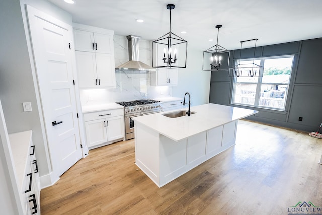 kitchen with sink, wall chimney range hood, high end stove, an island with sink, and decorative light fixtures